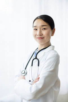 Asian Female Doctor in White Coat Smiling with Arms Crossed by Bright Window