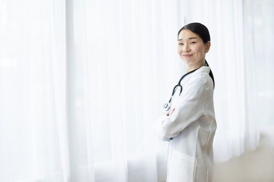 Asian Female Doctor in White Coat Smiling with Arms Crossed by Bright Window