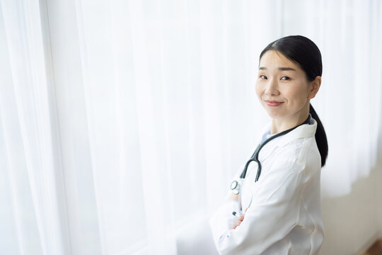 Asian Female Doctor in White Coat Smiling with Arms Crossed by Bright Window