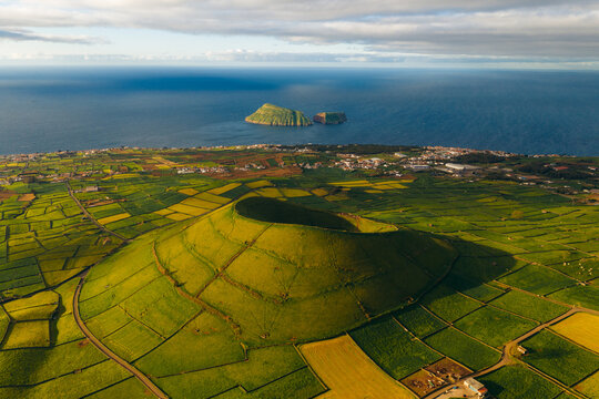 Aerial view of Pico Dona Joana geodesico volcanic cone surrounded by green patchwork fields and the Atlantic Ocean in Feteira, Acores, Portugal.