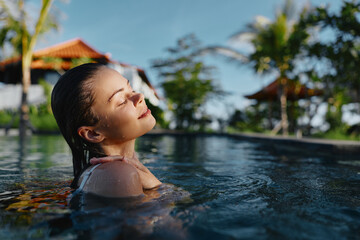Fototapeta premium A woman enjoys warm sunlight as she relaxes in a bright outdoor pool, water caressing her shoulders. Calm, refreshing atmosphere, leisure and wellness tied to summer self-care and tranquil moments.