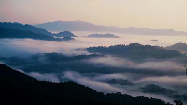 High-angle view of a moving sea of clouds flowing through mountain valleys. Dense mist and fog settle between dark silhouetted peaks during a soft, pastel morning dawn.