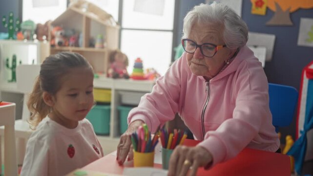 Elderly woman guiding a child's drawing, pointing at paper with colored pencils on table inside a classroom building; nurturing bond learning.