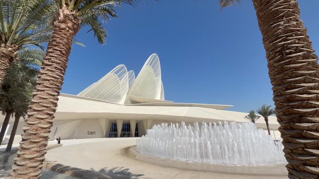 Wide cinematic exterior shot of the Zayed National Museum in Abu Dhabi framed by palm trees and fountain with futuristic white architecture under clear blue sky.