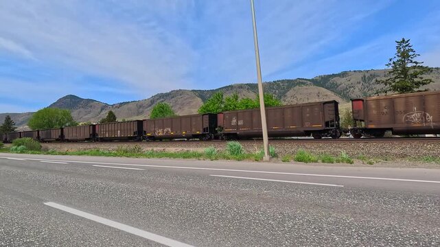 Freight train moving parallel to a main road or highway, with mountains and blue sky in the background.