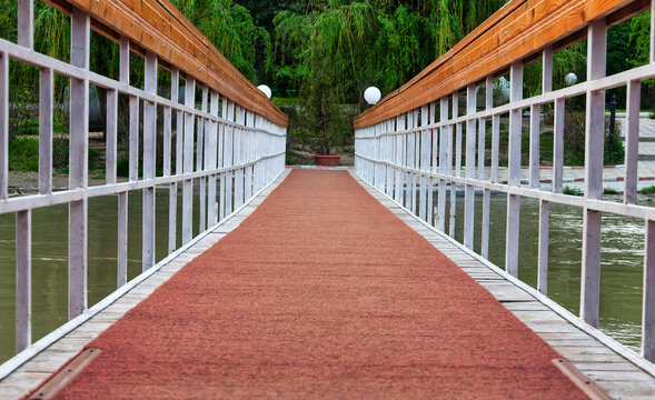 Wooden Footbridge with White Railings Extending Across a Calm River leading toward a green park shore.