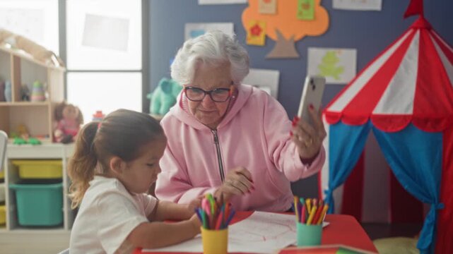 Senior woman shows smartphone while little girl covers ears with hand at classroom building table with crayons and toys; joyful bonding play.