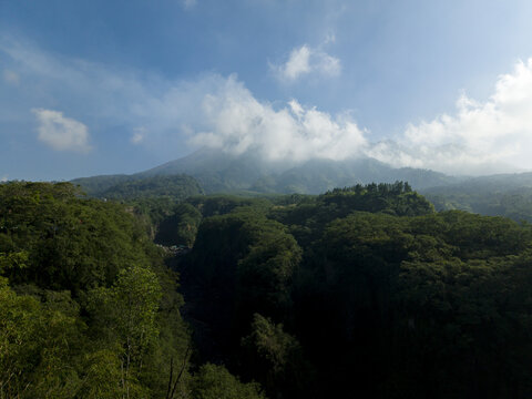 Aerial view of the Tambang Pasir Merapi Kali Gendol Sand Mine surrounded by dense tropical forest with Mount Merapi under a cloudy sky Kecamatan Cangkringan, Daerah Istimewa Yogya, Indonesia.