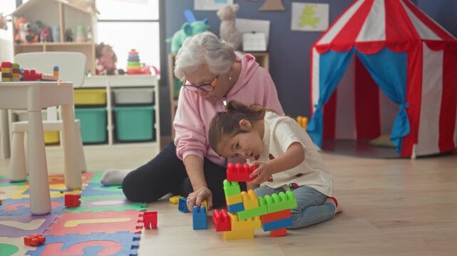 Elderly woman helps little girl stack colorful blocks on foam mat in a playroom with toy tent and storage bins; bonding play.
