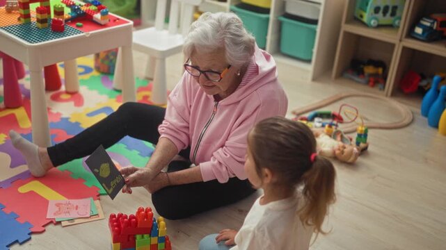 Woman holding and showing a card to a little girl on a playroom floor, pointing at the flashcard while seated; gentle learning warmth.