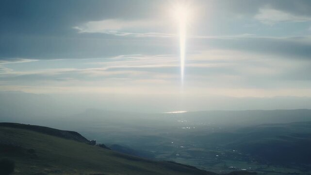 Bright sun pillar rising vertically from the horizon into the hazy sky, illuminating a vast mountain valley and distant body of water under a cloudy meteorological optical phenomenon