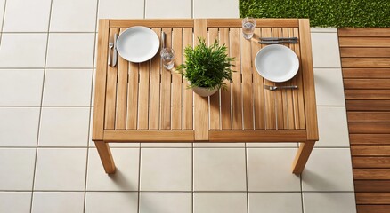 Naklejka premium Top down view of small wooden outdoor table set for two, complete with plates, cutlery, and small potted plant, situated on tiled patio next to wooden deck and grass.