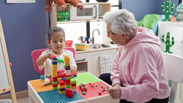 Senior woman reaches to stack colorful toy blocks with little girl at a small play table in a building; tenderness intergenerational bonding.