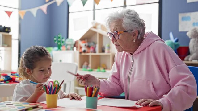 Senior woman helps little girl draw with colored pencils and paper at a preschool classroom table, guiding her hand and pointing to shapes; tender learning.