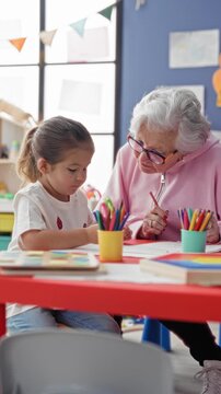 Senior woman guides little girl drawing, holding pencil and pointing at paper at classroom table in preschool; intergenerational learning warmth.