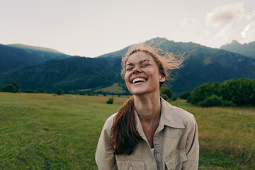 Fototapeta premium Bright joyful young woman laughs in a sunlit field with distant mountains, hair blown by the breeze, wearing a casual beige jacket and relaxed pose