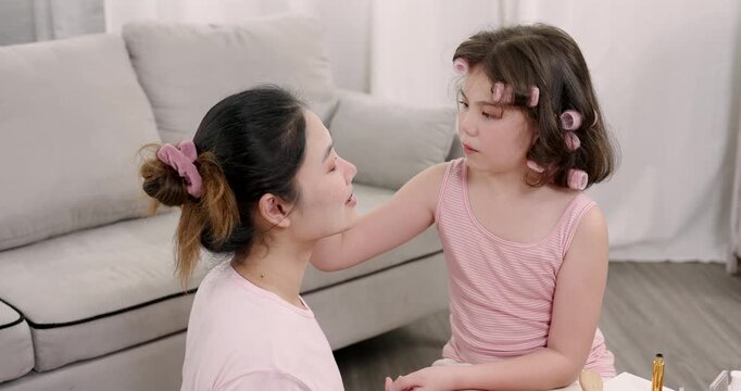 Asian girl gently applies mascara on single mother face during beauty learning time while sitting on floor together smiling with cosmetic tools in warm bonding family moment
