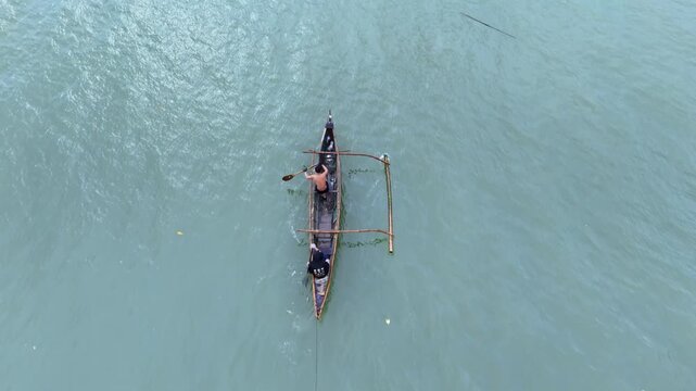 Top view of traditional wooden outrigger canoe paddling across calm turquoise ocean water with man.