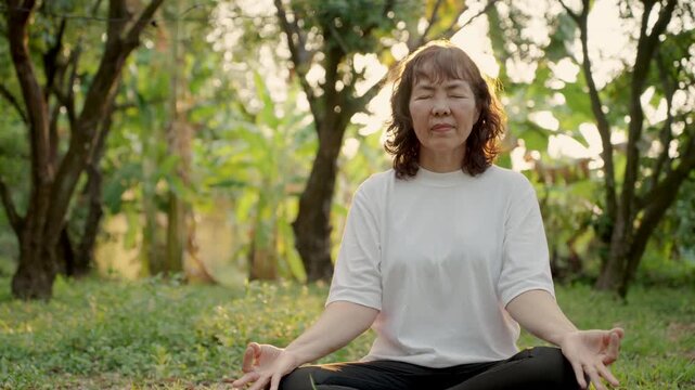 Happy senior woman meditating on yoga mat in green park, enjoying peaceful retirement lifestyle. Wellness, mindfulness, healthy aging and relaxation in nature concept.
