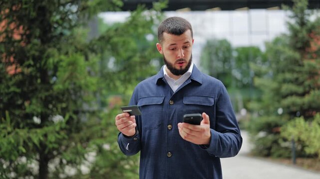 Young man baffled by credit card and smartphone outside. He faces declined payment, blocked account or banking app error. Focus on financial stress, money problems and technical tech glitches.