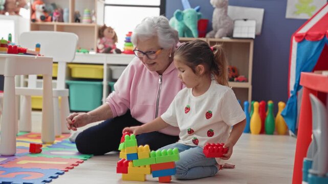 Woman building toy blocks with little girl on foam mat in a preschool playroom, sitting on floor and stacking colorful bricks; nurturing connection.