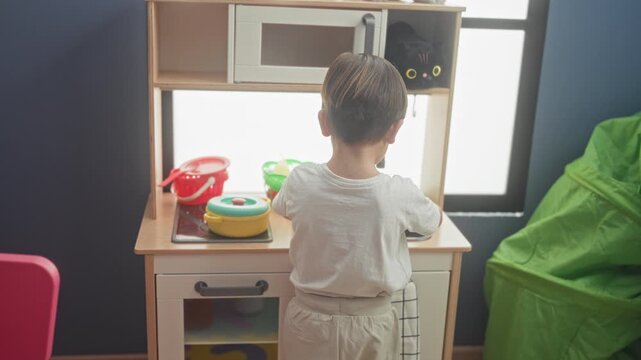 Boy stirs a yellow toy pot with a red spoon on a wooden play kitchen counter in a building; playful curiosity.