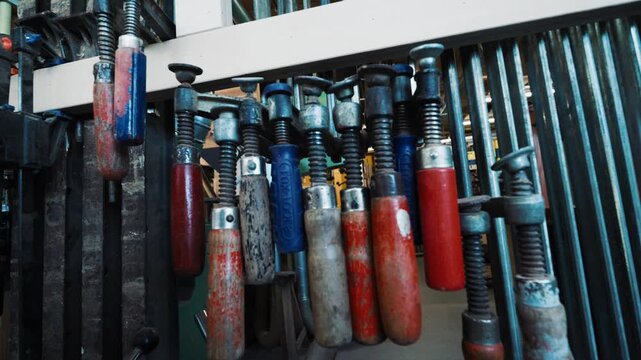 Rack of clamps and hand tools hanging on a wall inside a woodworking workshop.