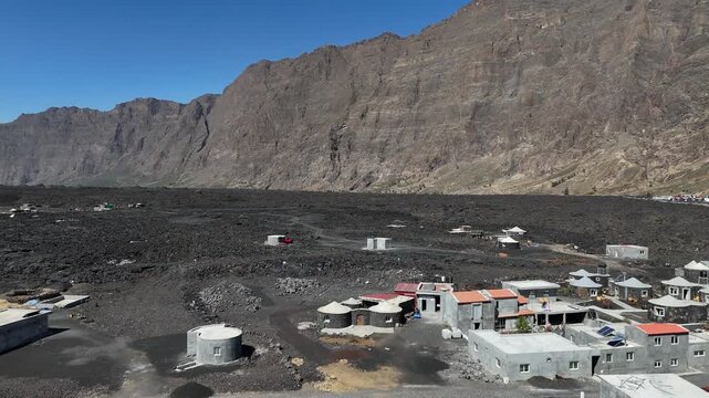 Aerial drone shot of the Ch&atilde; das Caldeiras village and traditional stone houses situated on black volcanic soil beneath the massive Bordeira cliff wall in Fogo National Park, Cabo Verde