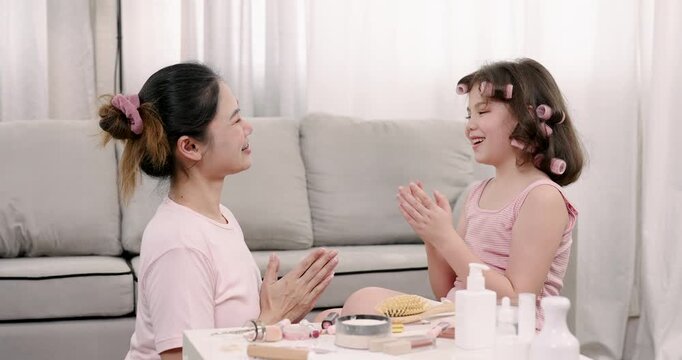 Multiracial girl holding mascara tube and wand while preparing to apply on Asian woman seated nearby, surrounded by makeup tools, showing focus and pretend makeup activity
