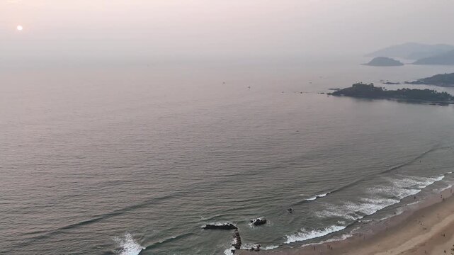 Wide cinematic aerial of the sunset coast in Goa showcasing textured ocean waters, subtle shoreline waves, and warm evening haze blending seamlessly into the horizon.