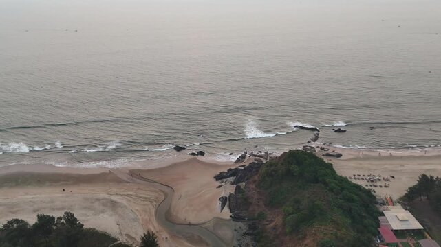 Wide cinematic aerial of the sunset coast in Goa, rugged tropical coastline where waves crash against scattered rocks, while a narrow stream flows across the sandy beach beside a lush green cliff edge