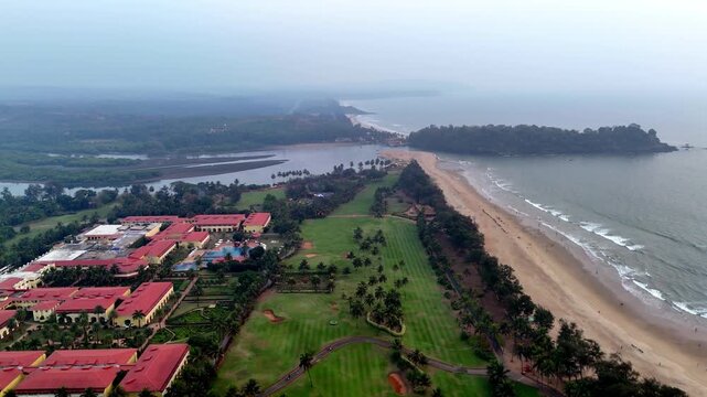 Cinematic aerial view of Canacona showcasing a luxury coastal resort beside a long sandy beach, with lush green lawns, palm-lined landscapes, and calm ocean waves stretching into the horizon.