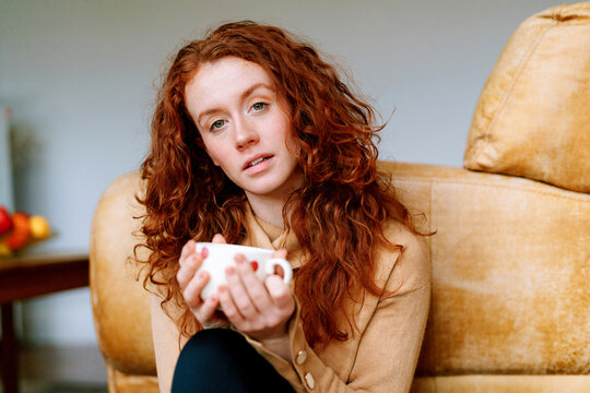 Young woman sits on couch holding a cup while looking thoughtfully at the camera in a cozy living room setting