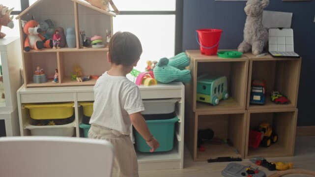 Boy reaches with hand to grab toy bus and red bucket from wooden cubby shelf while standing in a child's play area inside a building; curious imagination play.