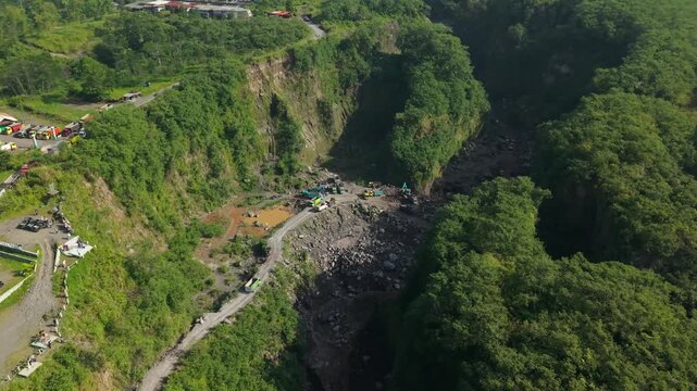 Aerial view of Tambang Pasir Merapi Kali Gendol sand mine with trucks and excavators surrounded by green cliffs in Kecamatan Cangkringan, Daerah Istimewa Yogya, Indonesia.