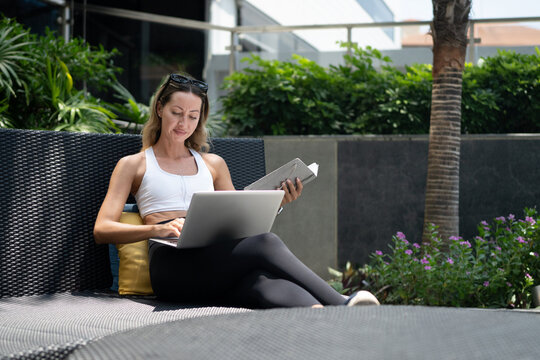 Active Woman Working Outdoors on Laptop with Notebook in Sporty Athleisure Wear at Modern Patio