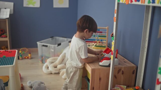 Boy presses buttons on a toy cash register and slides abacus beads at a wooden table in a building; quiet concentration.