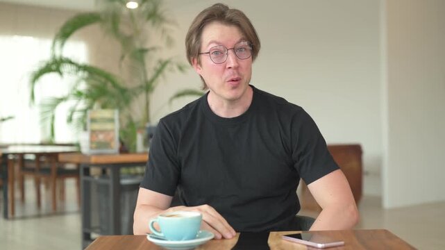 Portrait of Scandinavian Northern European man sitting in coffee shop cafe restaurant vlogging and talking to camera happily