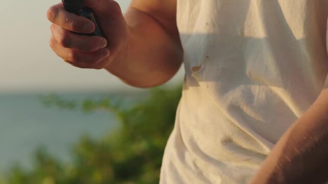 A slow motion close up of a white male spraying perfume on their muscled, veiny wrist with the ocean in the background