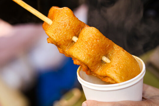 A classic Korean fish cake (eomuk or odeng) threaded onto a long wooden skewer. The fish cake is hot, resting across the top of a white paper cup filled with savory broth.