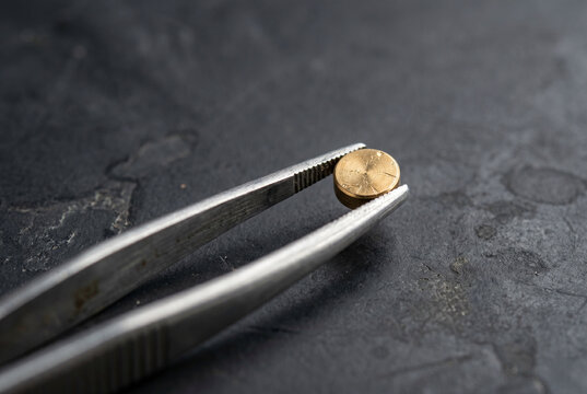 Stainless steel forceps holding a miniature 1g brass weight against a dark anthracite background. The shot emphasizes the delicate nature of high-end scientific calibration and handling.