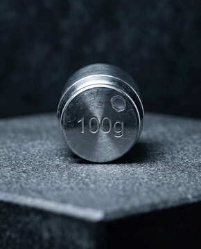 Vertical macro of a 100g stainless steel weight. The top-down lighting highlights industrial cleaning marks and the cold, unyielding beauty of high-grade laboratory equipment.