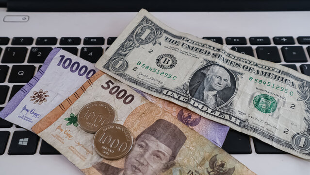 Comparison and exchange rate of US dollar and Indonesian rupiah banknotes, with a laptop in the background.