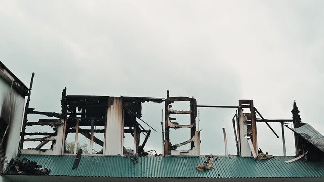 Smoke billows from the remains of a burned structure. Firefighters are present, working to extinguish the remaining flames and secure the area
