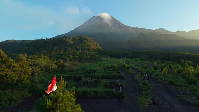 Aerial view of Bunker Kaliadem Merapi and Mount Merapi with pine trees and people walking on volcanic soil under a clear sky in Kecamatan Pakem, Yogyakarta, Indonesia.