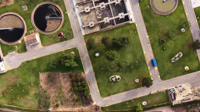 Top-down view of an urban wastewater treatment plant featuring circular clarifier tanks, rectangular aeration basins, green landscaped grounds and internal road network