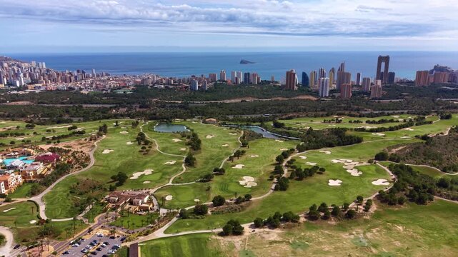 Panoramic view of Benidorm golf course with lush fairways, sand bunkers and water hazards, city skyline and Mediterranean Sea visible in background