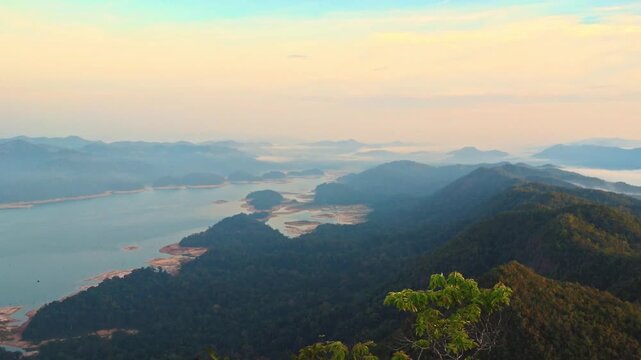 High-angle panoramic pan from a mountain peak. A vast lake and river system meet a thick sea of clouds nestled between silhouetted ridges under a soft morning dawn sky.