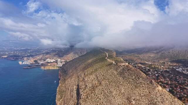 Morro de Toix viewpoint over Calpe, Spain, revealing rocky coastal cliffs, Mediterranean Sea, low clouds over mountain ridge and seaside town below