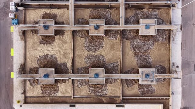 Overhead view of aeration basins with activated sludge at an urban wastewater treatment plant, showing turbulent brown water and mechanical aerators in concrete tanks
